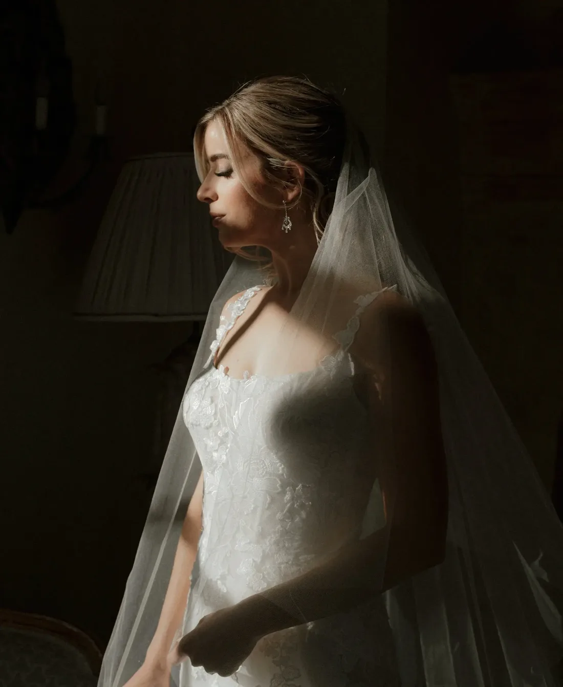 A bride in soft window light, her veil falling over her shoulder, on the morning of her wedding